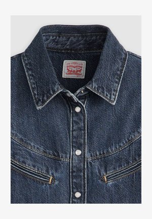 Tummansininen farkkutakki, jossa on kärjellinen kaulus, kaksi rintataskua ja nappikiinnitys, jossa on valkoiset napit ja erottuva keltainen ompelu.