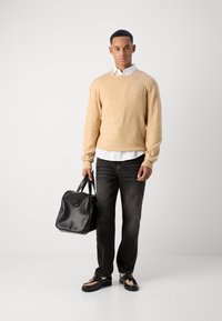 Helles beiges Stricksweatshirt über einem weißen Hemd mit Kragen, schwarze Jeans und schwarze Tasche mit Griffen; die Schuhe haben ein strukturiertes Design.