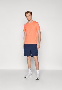 Herre træningsoutfit bestående af en klar orange Nike t-shirt og marineblå shorts med et hvidt Nike-logo, parret med hvide sneakers.