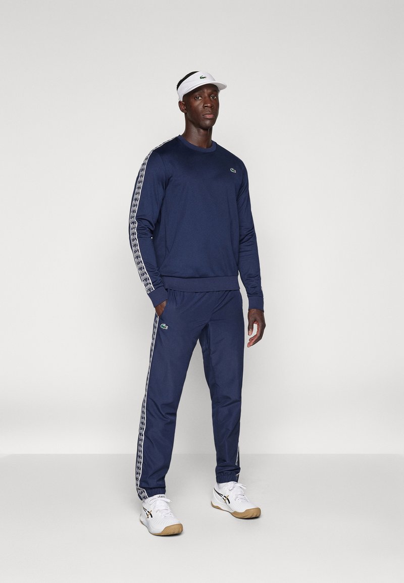 Uomo in piedi che indossa una tuta sportiva blu navy con strisce bianche a fantasia e un visiera bianca, abbinata a sneakers bianche, contro uno sfondo semplice.