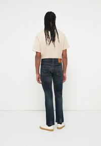 Camiseta color beige claro, jeans de mezclilla oscuros con corte recto y zapatillas blancas con suela beige. Presenta un parche de cuero en la parte trasera de la cinturilla.