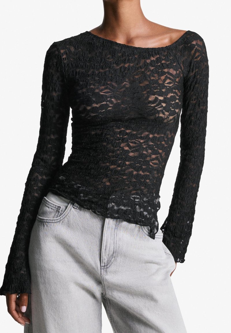 Femme portant un haut noir en dentelle à manches longues ajusté, associé à un jean en denim gris clair taille haute, sur fond blanc.
