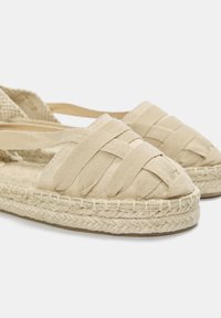Béžové espadrilky s tkaným textilním svrškem, juteovou podrážkou a kulatou špičkou. Měkká plátěná textura s elastickými popruhy pro bezpečné nošení.