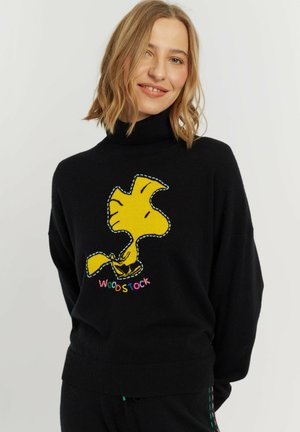 Femme souriante aux cheveux blonds porte un pull à col roulé noir avec un grand personnage Woodstock jaune et un texte multicolore à l'avant.