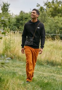 Felpa nera con logo, pantaloni arancioni e una piccola borsa a tracolla nera. Uomo che cammina in un'area erbosa, con scarpe da ginnastica. Sfondo nuvoloso.