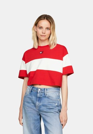 Donna che indossa una t-shirt corta rossa con una larga striscia orizzontale bianca e jeans a vita alta azzurri, in piedi contro uno sfondo neutro.