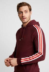 Burgundy hoodie with a drawstring and kangaroo pocket. Features red, white, and yellow stripes on the sleeves. Soft fabric texture.