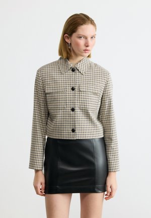 Vrouw draagt een beige geruit jasje met zwarte knopen en een zwarte leren minirok, staand tegen een effen achtergrond.