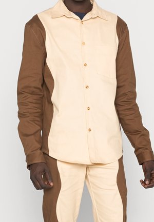 Camicia a bottoni realizzata in cotone con tessuto beige e marrone a contrasto, tasca sul petto e orlo curvo, con dettagli di cucitura a vista.