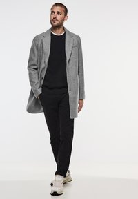 Grå ullblandad kappa med skräddarsydd design, enfärgad knäppning framtill och slagkrage, bärs över en svart tröja och svarta byxor, tillsammans med sneakers.