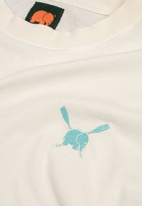 Biely bavlnený tričko s svetlým modrým vyšívaným dizajnom slona s králičími ušami a zelenou nášivkou s oranžovým logom slona.