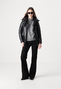 Giacca a puffo nera con finitura lucida, maglione grigio a collo alto, pantaloni neri a zampa d'elefante e occhiali da sole neri oversize. Ambientato su uno sfondo neutro.