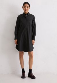 Femme portant une chemise robe noire à manches longues avec des chaussettes noires et des mocassins marron foncé, se tenant contre un mur blanc.