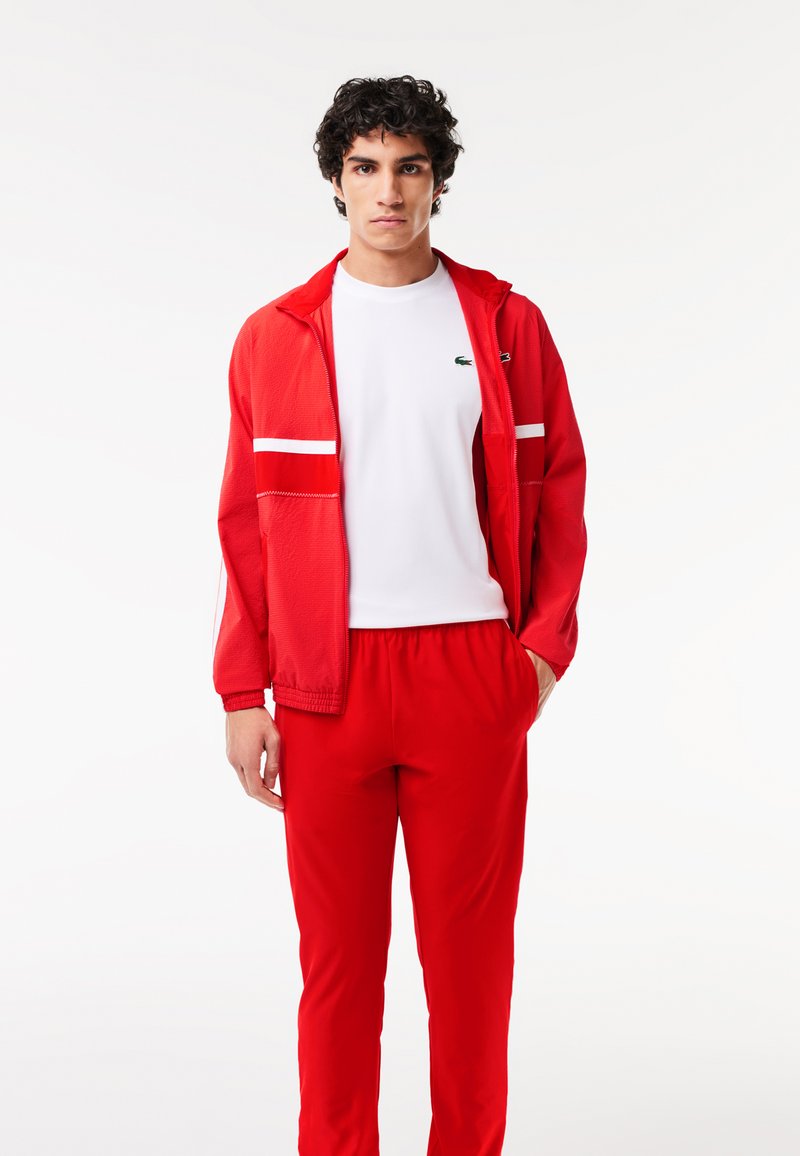Ensemble de sport rouge comprenant une veste zippée avec des rayures blanches et un pantalon assorti, accompagné d'un t-shirt blanc uni. Texture lisse.