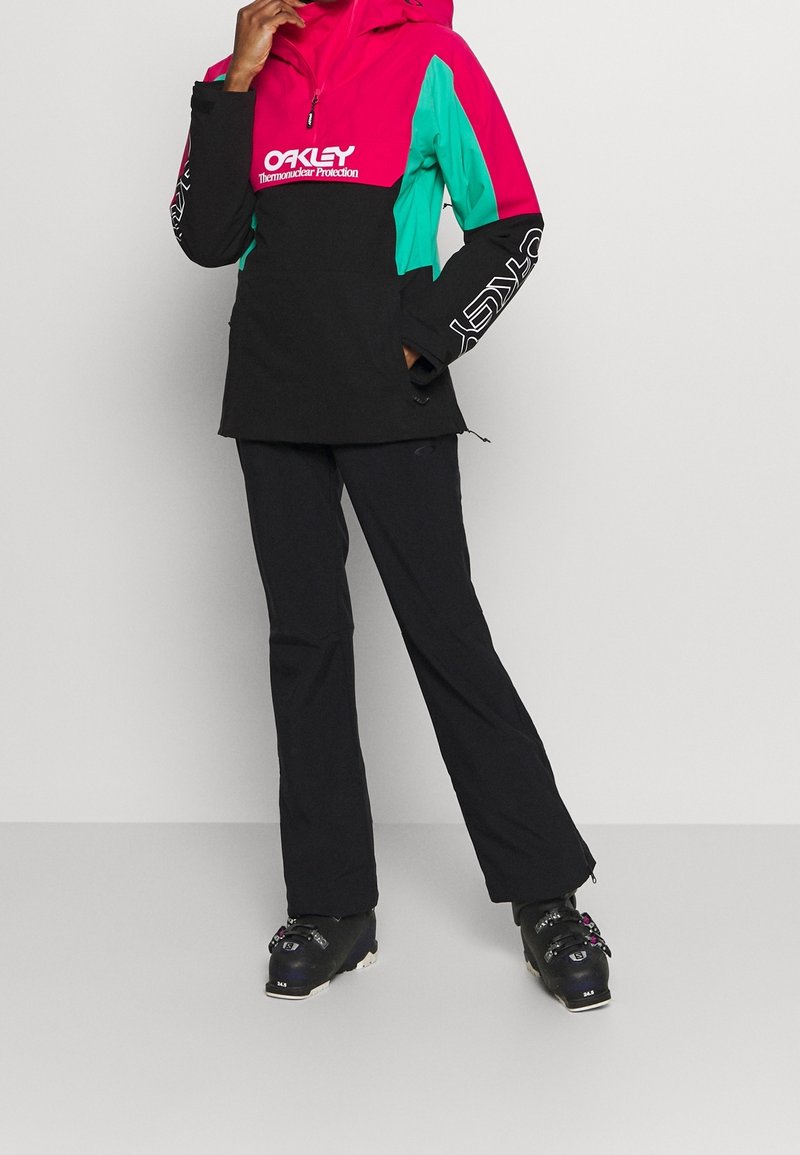 Personne portant un pantalon de ski noir, des chaussures de ski noires et une veste de ski color-block rose, bleu sarcelle et noir avec la marque Oakley.