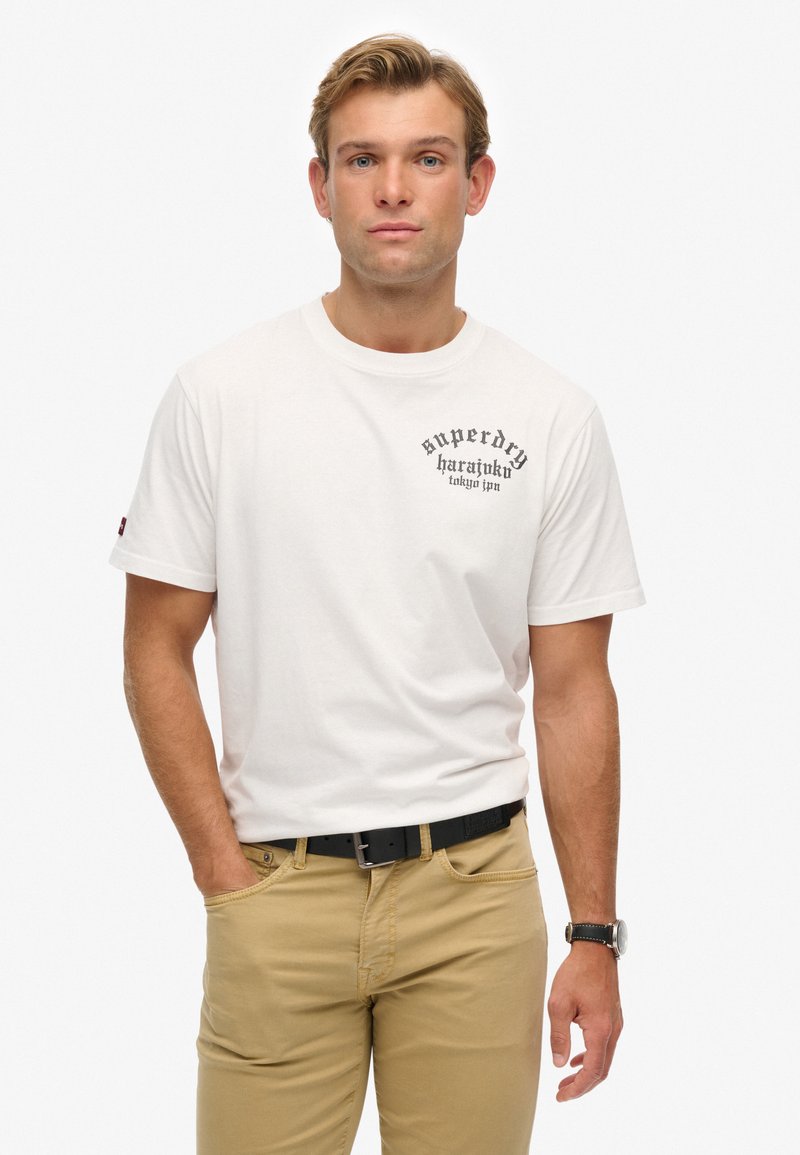 Camiseta de algodón blanca con estampado gráfico negro en el pecho, mangas cortas y cuello redondo. Combinada con pantalones caqui y un cinturón negro.