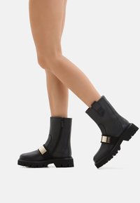 Legs wearing black mid-calf boots with chunky soles, side zippers, and gold rectangular accents, posed against a white background.