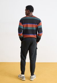 Csíkos, hosszú ujjú ing zöld, barna és kék csíkokkal, fekete cargo nadrággal és szürke sportcipővel párosítva. Texturált anyag és laza szabás.