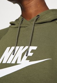 Olivgrön hoodie i strukturerad tyg, med en vit Nike-logotyp, dragsko med svarta spetsar och en klassisk design.