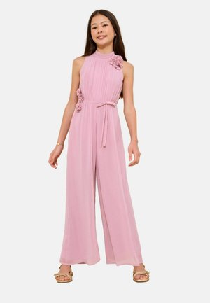 Noor tüdruk, kes kannab roosat varrukateta laiade säärtega kombinesooni lillemustriga ja vööga, seisab ja naeratab kuldsete sandaalidega.