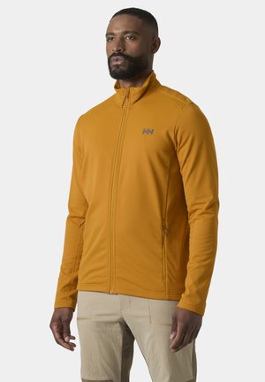 Mężczyzna w musztardowożółtej bluzie z suwakiem i logo HH oraz beżowych spodniach, stojący na tle jednolitego jasnego tła.