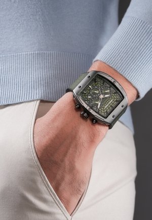 Muñeca con reloj cronógrafo verde oliva con esfera de patrón geométrico, llevado sobre un suéter azul claro y pantalones beige con la mano en el bolsillo.