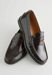 Mocasines de cuero marrón brillante con una textura suave, presentan una costura elevada y una tira decorativa sutil en el empeine. Suela de goma negra.