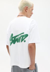 Witte t-shirt met korte mouwen en een groen graffiti-stijl logo op de achterkant. Gemaakt van een gestructureerde stof, met een relaxte pasvorm en een ronde zoom.