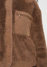 Veste beige texturée avec un extérieur doux et duveteux, une poche poitrine matelassée et une fermeture à boutons-pression. Détails en brun plus foncé.