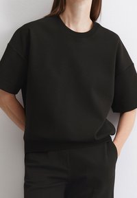 Černé tričko s kulatým výstřihem a krátkými rukávy, vyrobené z hladké směsi bavlny. Má volný střih a žebrovaný lem u výstřihu.