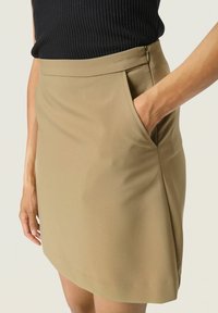 Beige szoknya sima textúrával, oldalzsebbel és diszkrét cipzár részlettel, egy modell kezén mutatva a derék közelében.