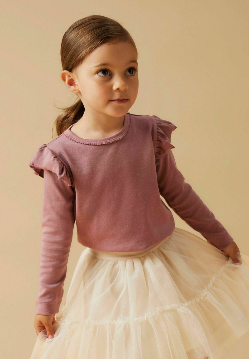 Jasnoróżowa koszula z długim rękawem z falbanami na ramionach, w połączeniu z warstwową spódnicą z kremowego tiulu. Gładka tekstura, casualowy styl.