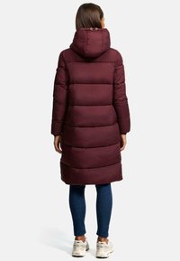 Bordeaux pufferjas met capuchon, voorzien van een horizontaal gestikt ontwerp, boven de knie lengte en verstelbare details. Draagt met blauwe jeans en sneakers.