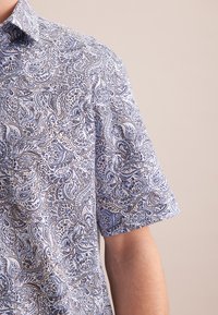 Koszula z krótkim rękawem w jasnoniebieskim kolorze z misternym wzorem paisley w ciemniejszym niebieskim. Zapinana na kołnierz z gładką, tkany teksturą.