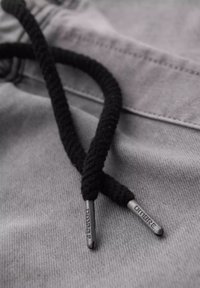 Cuerdas trenzadas negras con extremos de metal reposando sobre una tela gris, que presenta una textura suave y un sencillo detalle de costura en la parte superior.