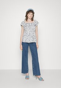 Top peplum floral branca com mangas franzidas, combinada com jeans de ganga azul de perna larga e sandálias de salto azuis. Fundo simples.