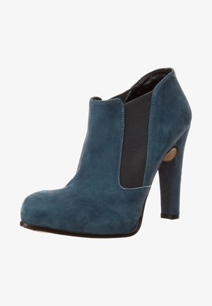 Sininen mokkanahkainen nilkkuri, jossa on korkea stiletto-korko, joustavat sivupaneelit ja pyöreä kärki. Suuret sileät pinnat, joissa on kontrastivärisiä mustia yksityiskohtia.