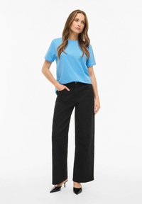 Niebieska bawełniana koszulka z okrągłym dekoltem i krótkimi rękawami, zestawiona z czarnymi szerokimi spodniami. Modelka nosi czarne szpilki z ostrym noskiem.