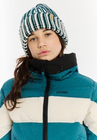 Pruhovaný puffer kabát v teal a krémovej farbe s vysokým golierom, čiernym zipsom a logom, doplnený pruhovanou pletenou čiapkou.