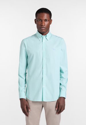 Mann trägt ein hellblau gestreiftes, langärmeliges Hemd mit Knöpfen und beige Hose, steht vor einem einfarbigen weißen Hintergrund.