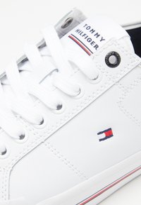 Weißer Ledersneaker mit flachen weißen Schnürsenkeln, versehen mit einem kleinen gestickten Logo in Rot, Weiß und Blau. Gummisohle mit gestreiften Akzenten.