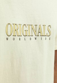 Camiseta de algodón de color beige claro con el texto en negrita "ORIGINALS" en negro y "WORLDWIDE" debajo en una fuente más pequeña. Diseño simple y casual.