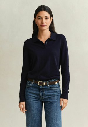 Jeune femme aux longs cheveux foncés porte un polo noir à manches longues rentré dans un jean bleu avec une ceinture marron, debout devant un fond uni.