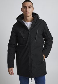 Parka neagră cu glugă, cu fermoare și buzunare laterale, realizată dintr-un material texturat; un design drept, cu sireturi ajustabile și un finisaj neted.