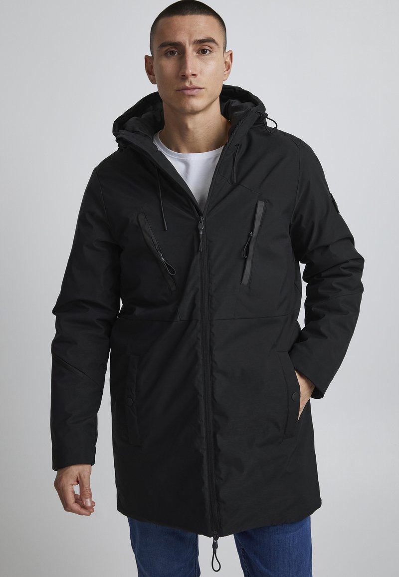 Parka neagră cu glugă, cu fermoare și buzunare laterale, realizată dintr-un material texturat; un design drept, cu sireturi ajustabile și un finisaj neted.