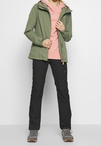 Lightweight green jacket with a zip front, paired with a pink hoodie and black pants. Gray athletic shoes complete the outdoor outfit.