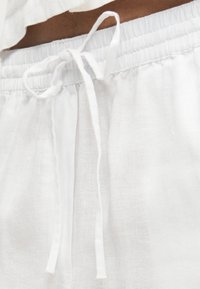 Λεπτομέρεια στη μέση λευκών λινών παντελονιών με λάστιχο και υφασμάτινο κορδόνι που δένει, φορεμένα με κοντό λευκό τοπ.