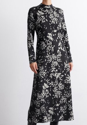 Robe midi noire à manches longues avec motif floral abstrait blanc, col montant, taille cintrée, portée par une personne debout sur un fond uni.