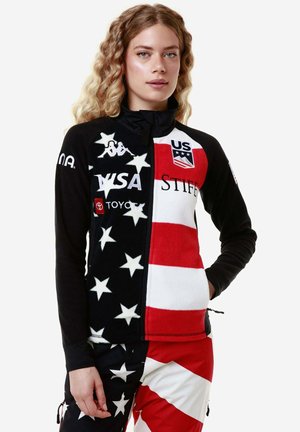Giacca in pile con design a spacco caratterizzata da stelle nere su sfondo bianco e righe rosse. Loghi sul petto e sulle maniche, colletto alto, chiusura con zip.