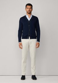 Cardigan lavorato a maglia blu navy con scollo a V e rifiniture bianche a contrasto, abbinato a pantaloni beige e scarpe scure, in piedi davanti a uno sfondo chiaro.